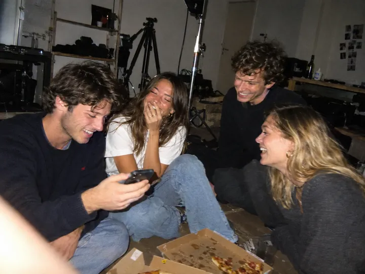 Four friends laughing on the floor of a messy studio late at night, pizza boxes at their feet