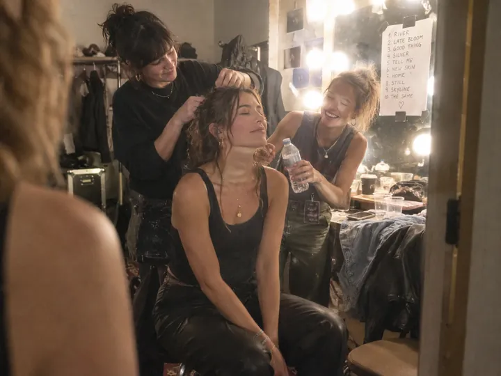A singer with her eyes closed in a small dressing room before a show, two friends fixing her hair and handing her water