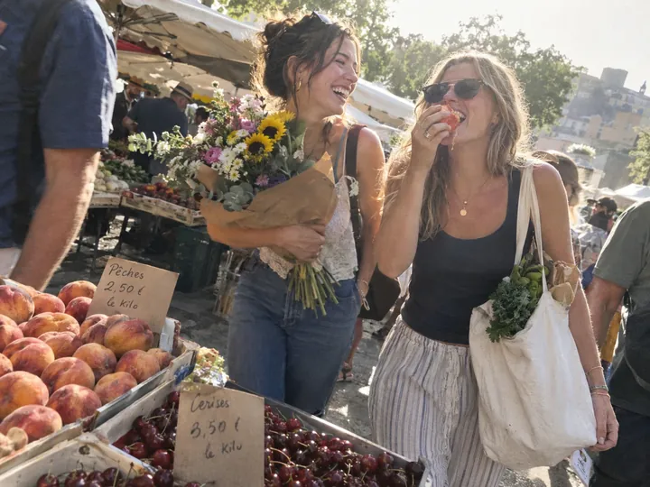 Two friends laughing at an outdoor market, one holding a bunch of flowers, the other biting into a peach